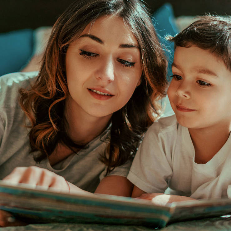 Woman and young boy reading a book together, lying on a bed in a cozy family setting