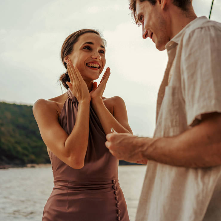 Man proposing on a yacht with woman in mauve dress, both smiling against scenic water and forested hills